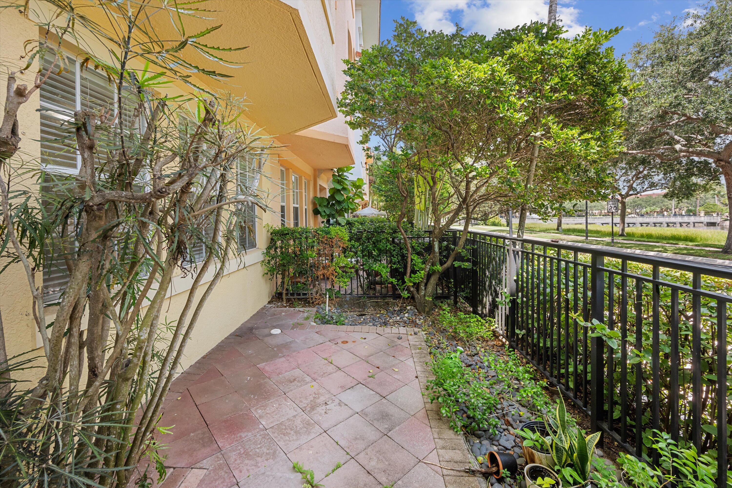 Paved patio beside a home, shaded by tropical trees with water visible beyond the railing.