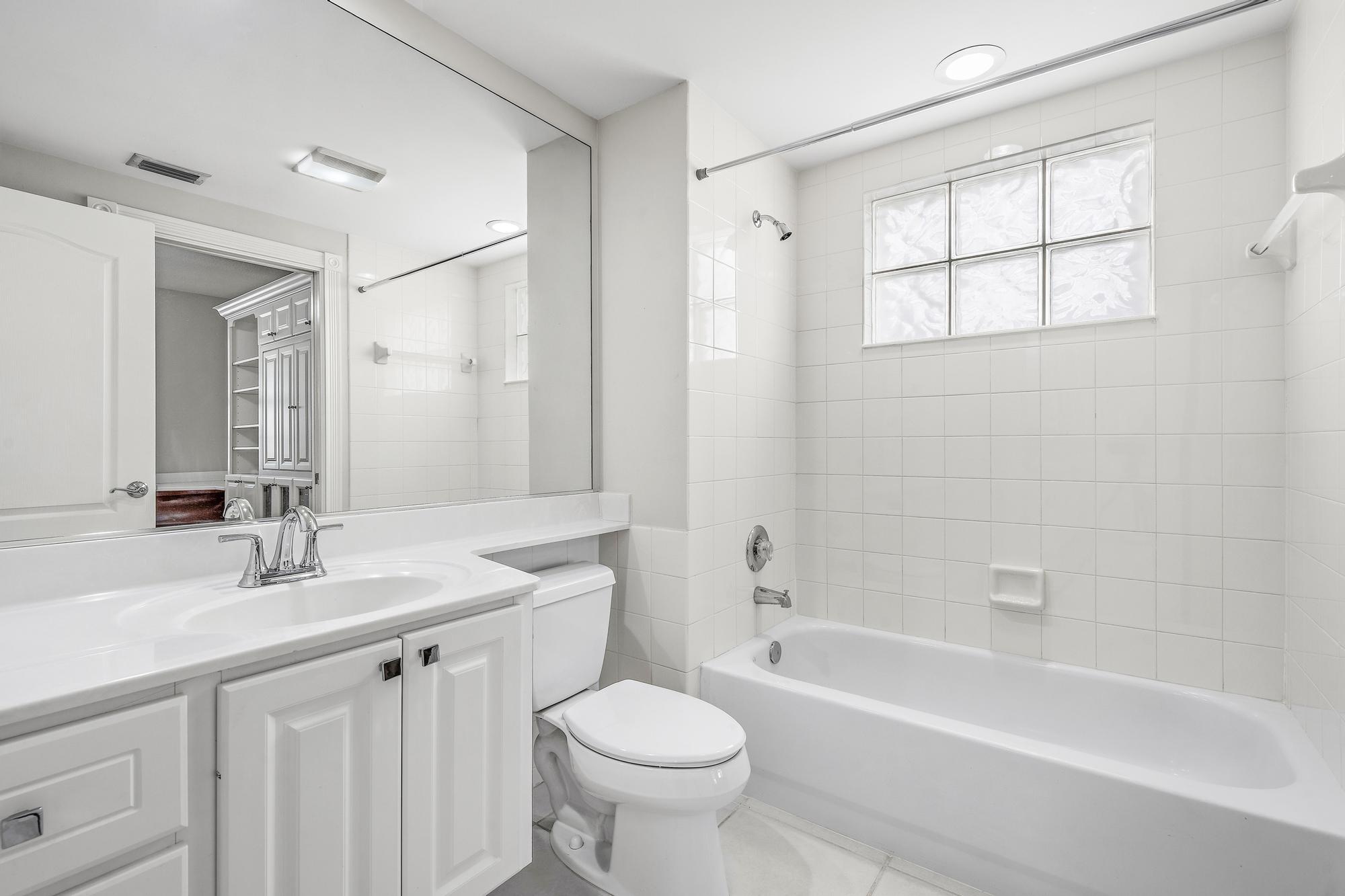 White bathroom with a tiled bathtub, glass-block window, and vanity beside the toilet.