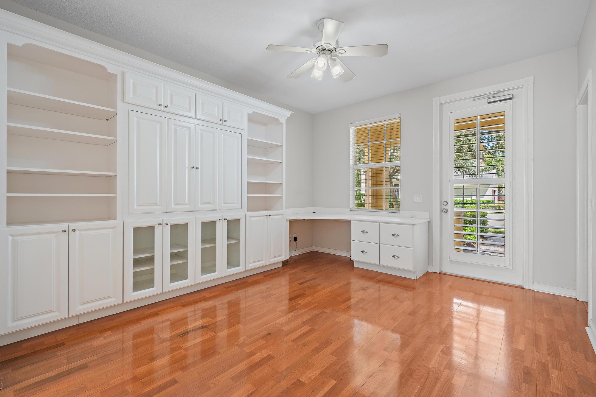Vacant home office with white built-in cabinets, desk, hardwood floors, and a shuttered glass door.