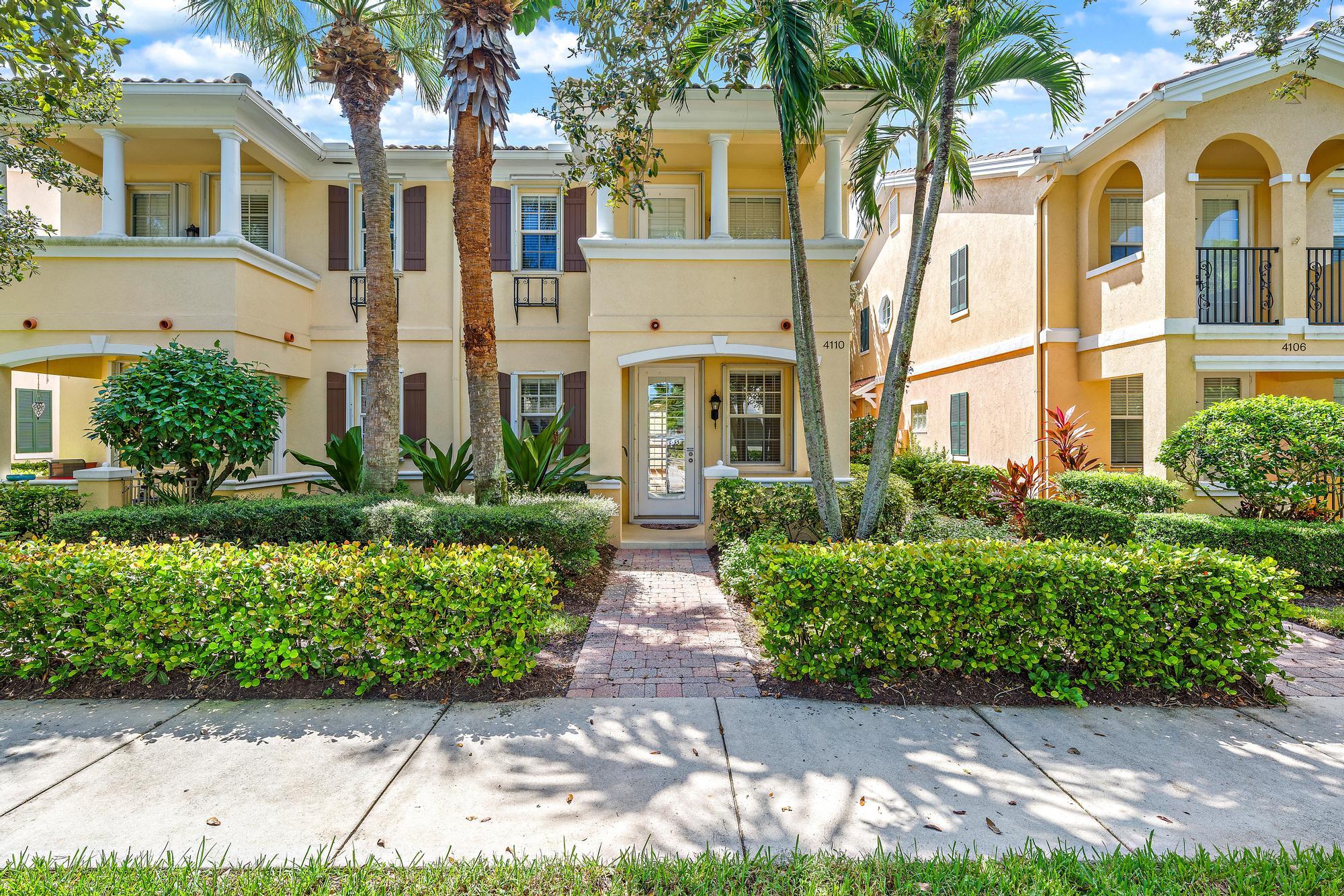 Two-story yellow townhouse with white columns, palms, and a brick entry path through trimmed hedges.