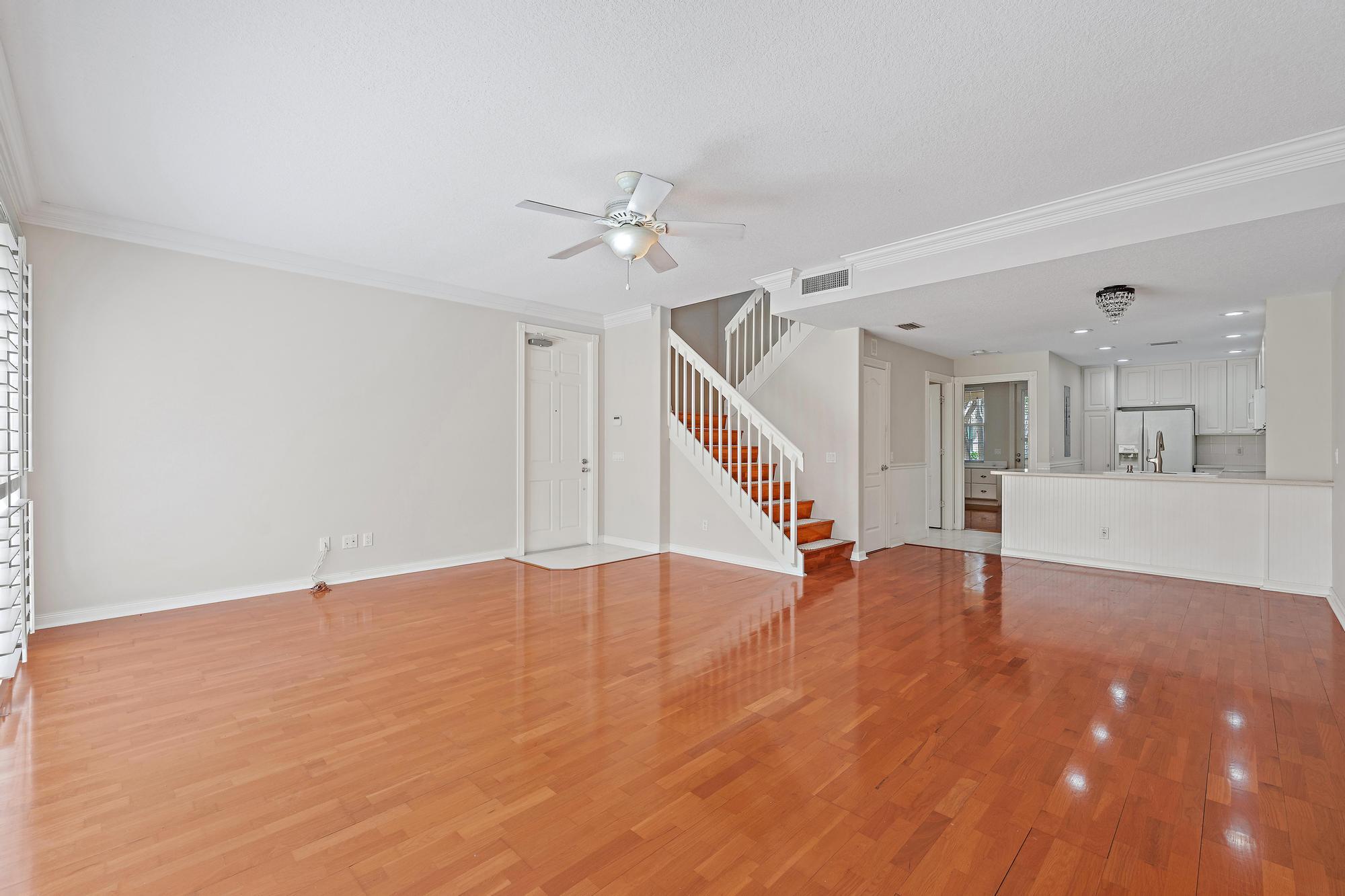 Vacant living room with polished hardwood floors, a white staircase, and an open kitchen.