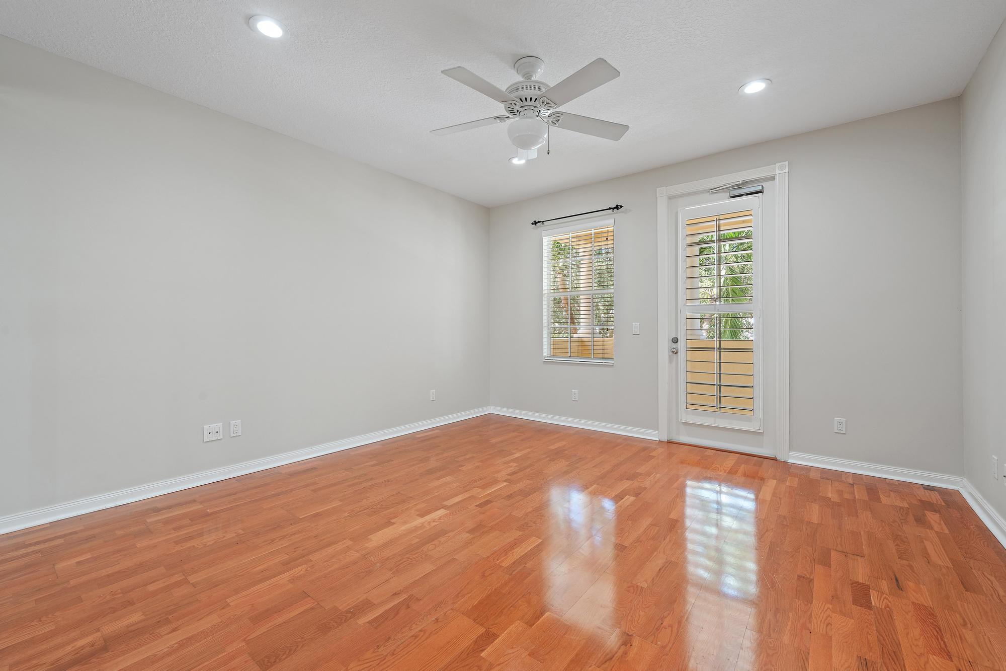 Empty room with polished hardwood floors, ceiling fan, and a glass door beside a window.