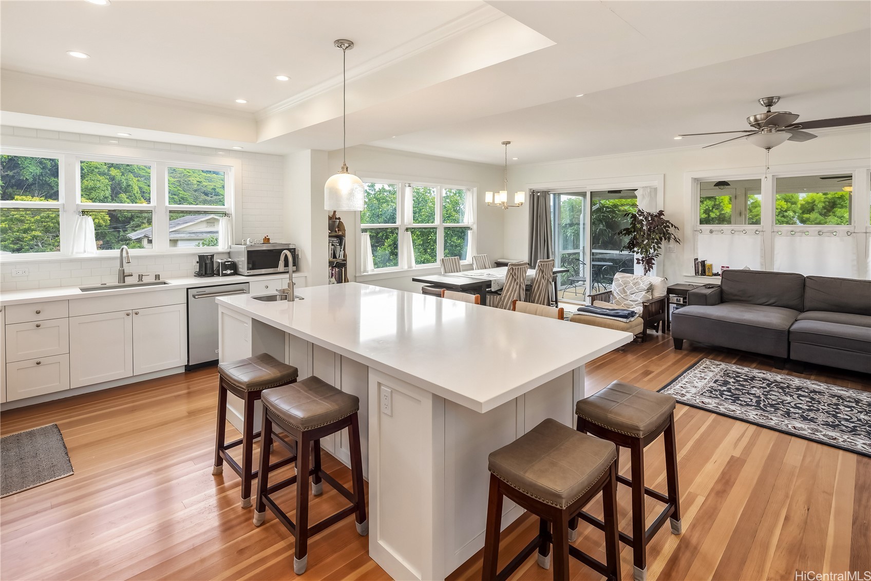 Open-plan kitchen with a large white island, hardwood floors, and windows overlooking greenery.