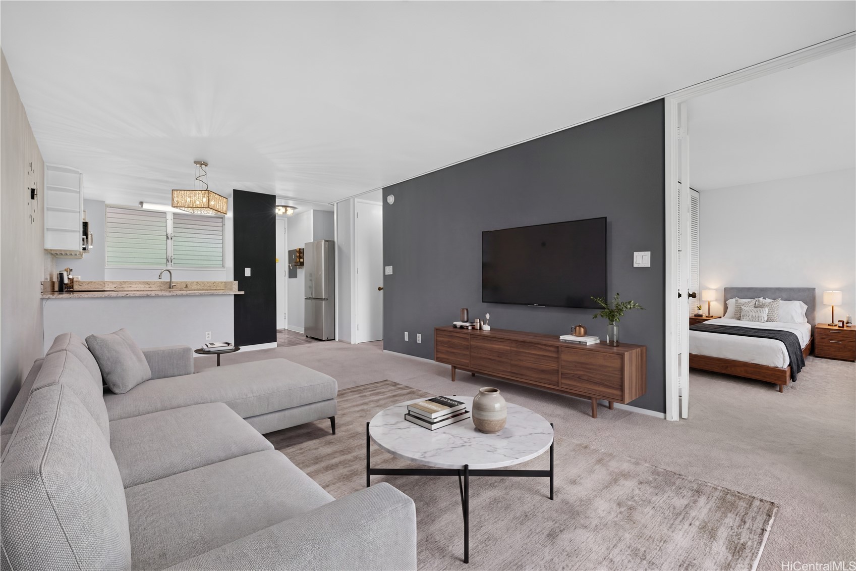Staged living room with a gray sectional, dark accent wall, open kitchen, and bedroom beyond.