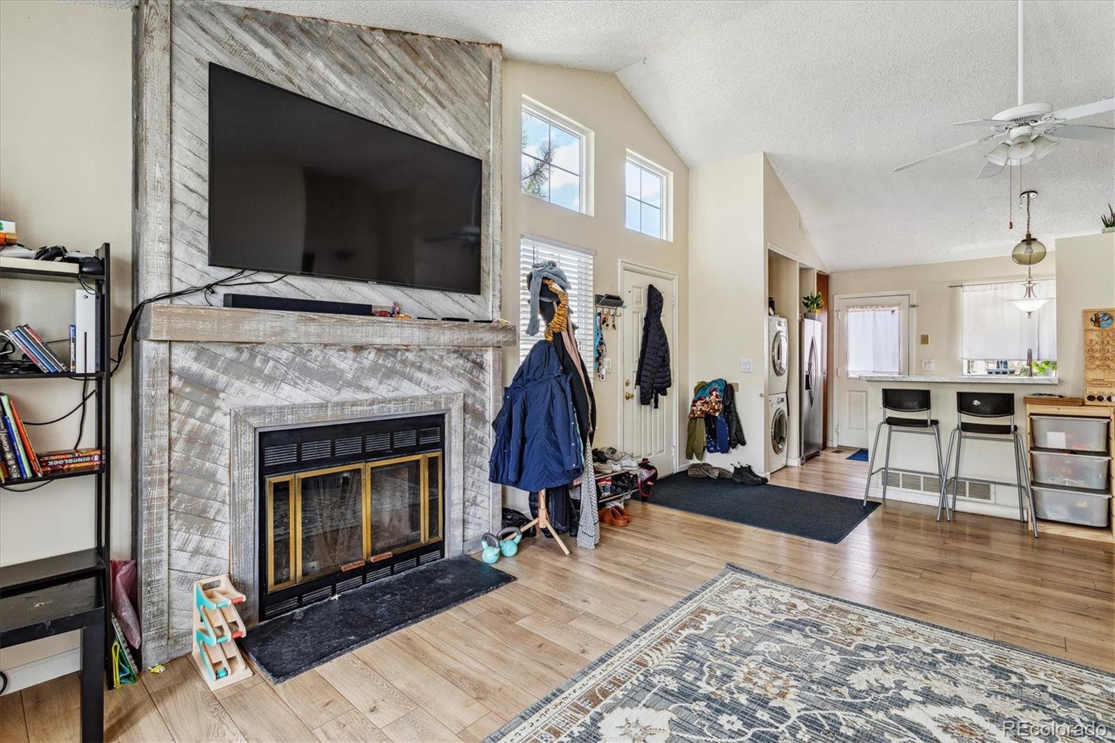 Open-plan living room with a large fireplace, mounted TV, vaulted ceiling, and kitchen counter.