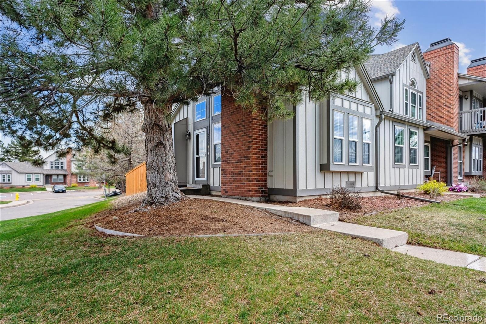 Townhouse exterior with gray siding, brick accents, a large pine tree, and a walkway to the entrance.