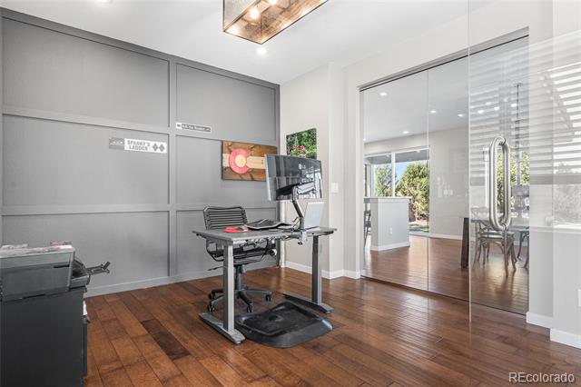 Home office with a desk and computer, gray paneled wall, glass doors, and hardwood floors.