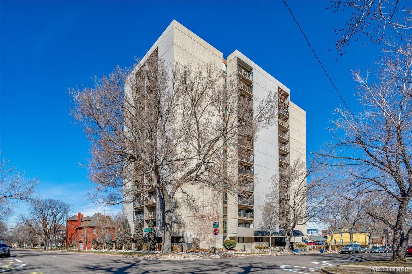 Beige condominium building with balconies behind leafless trees at a Denver street corner.