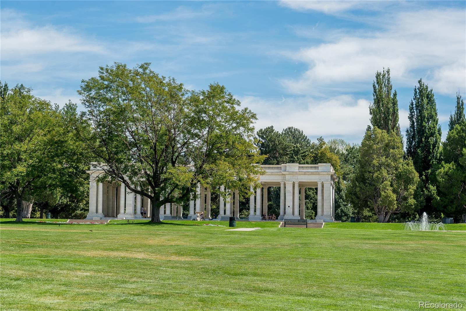 Park pavilion with white columns, mature trees, broad lawn, and a fountain.