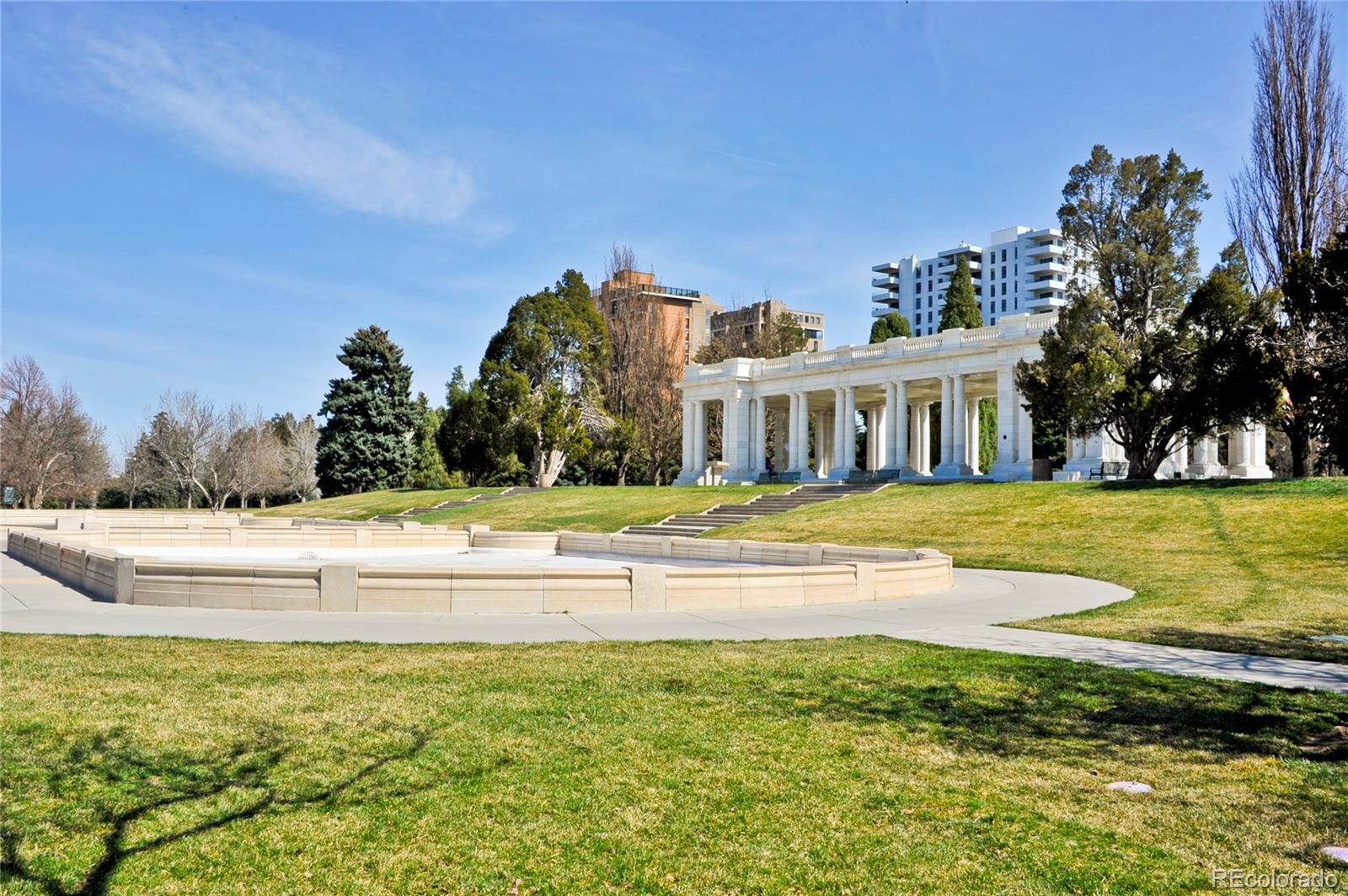 A white-columned pavilion stands beyond a circular stone basin in a grassy park.