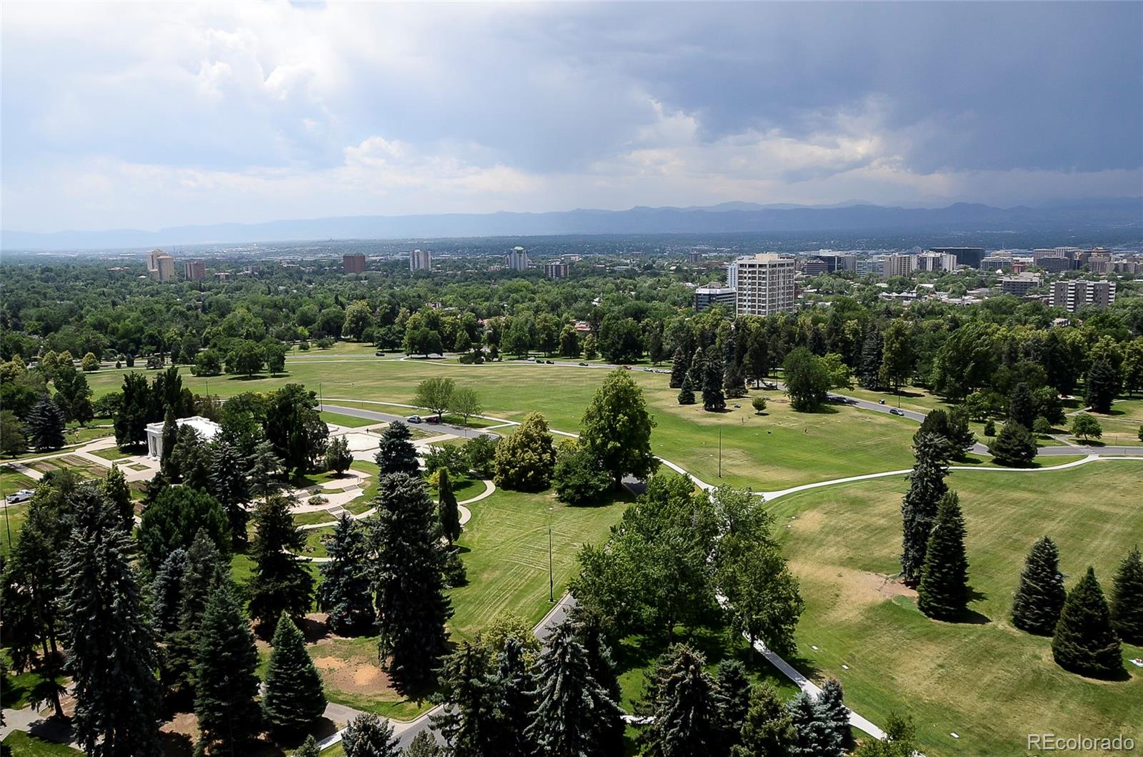 Panoramic view over a tree-filled Denver park toward the city skyline and distant mountains.