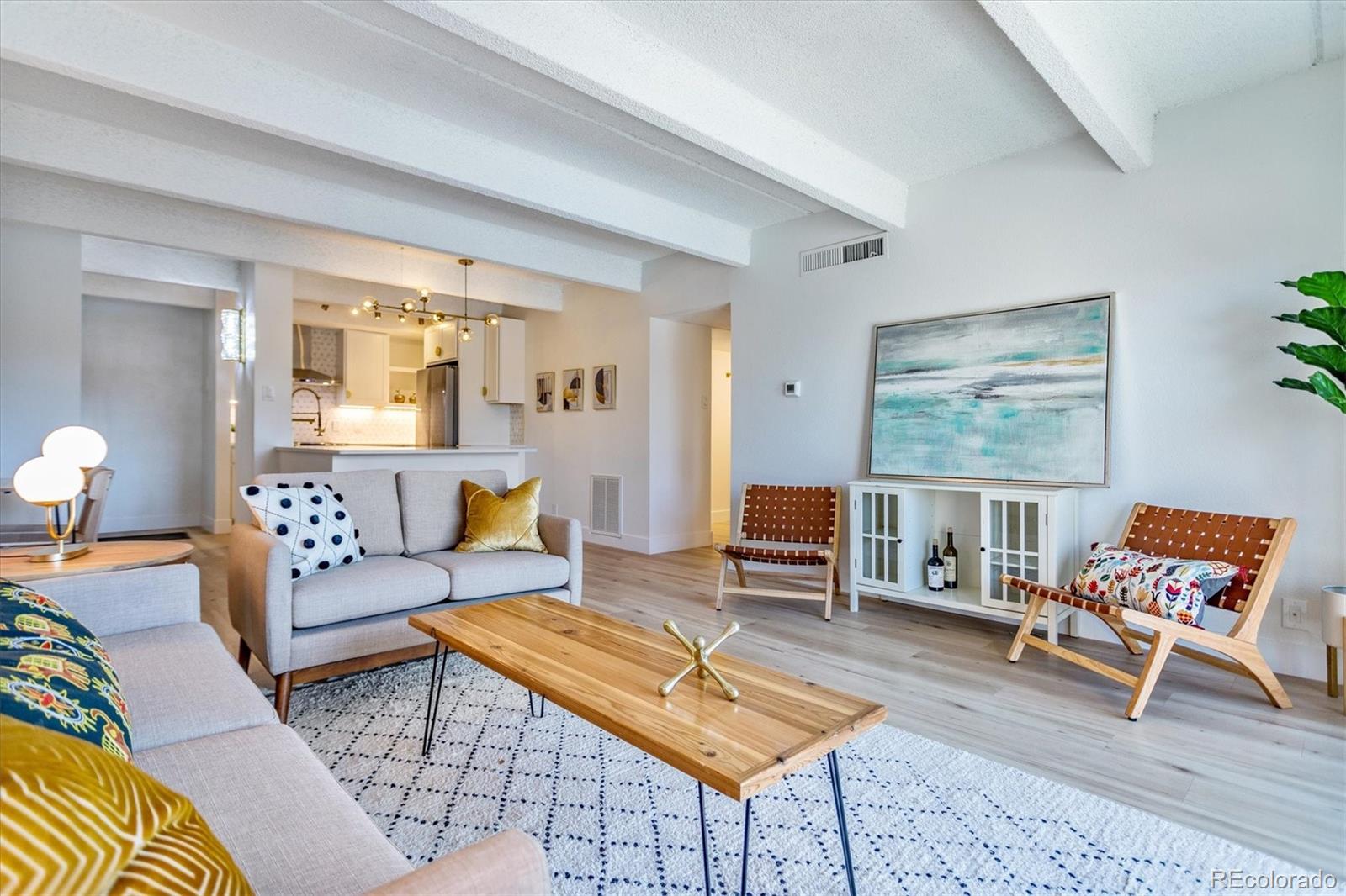 Open-plan living room with exposed ceiling beams, gray sofa, wooden chairs, and adjoining white kitchen.