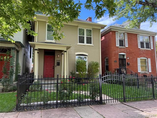 Two-story tan home with a red front door, landscaped garden, and black iron fence.
