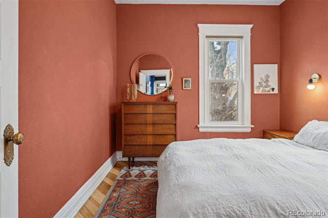 Bedroom with coral walls, a tall window, hardwood floors, and a wooden dresser beside a white bed.