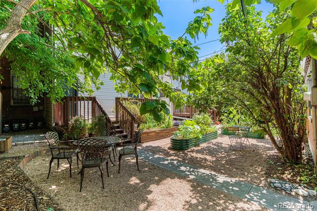 Shaded backyard patio with gravel seating area, mature trees, raised garden beds, and deck steps.