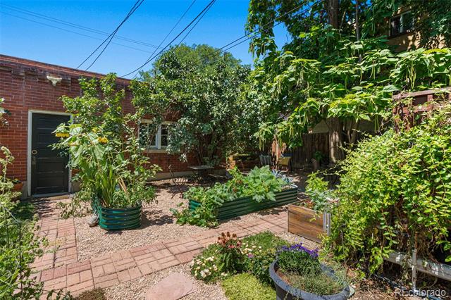 Brick-paved garden courtyard with raised beds and dense greenery beside a brick home.