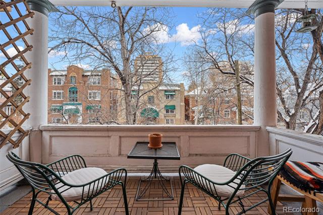Covered porch with black metal seating and a view of brick buildings through leafless trees.