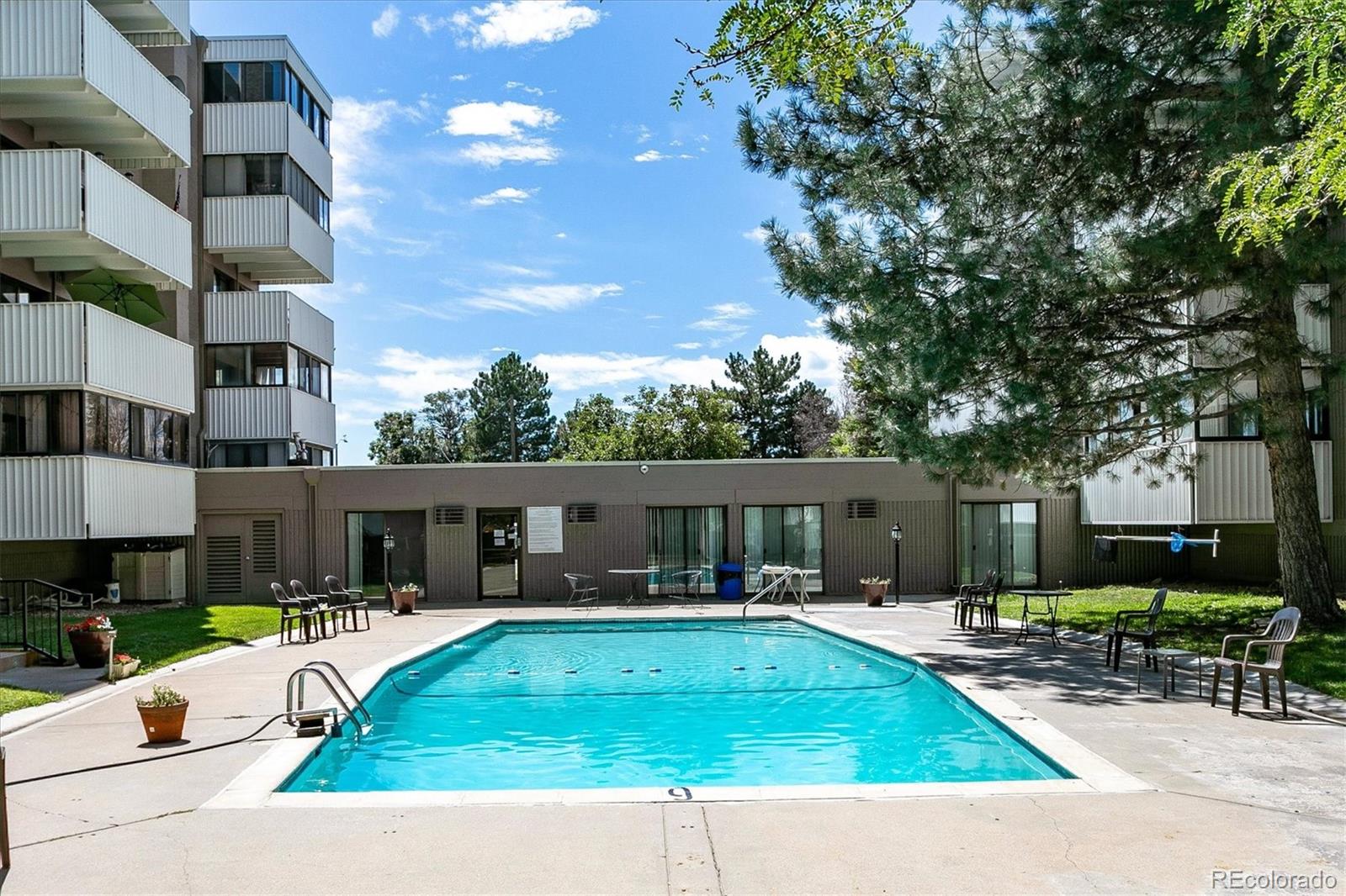 Outdoor pool beside an apartment building, with patio seating and mature trees.