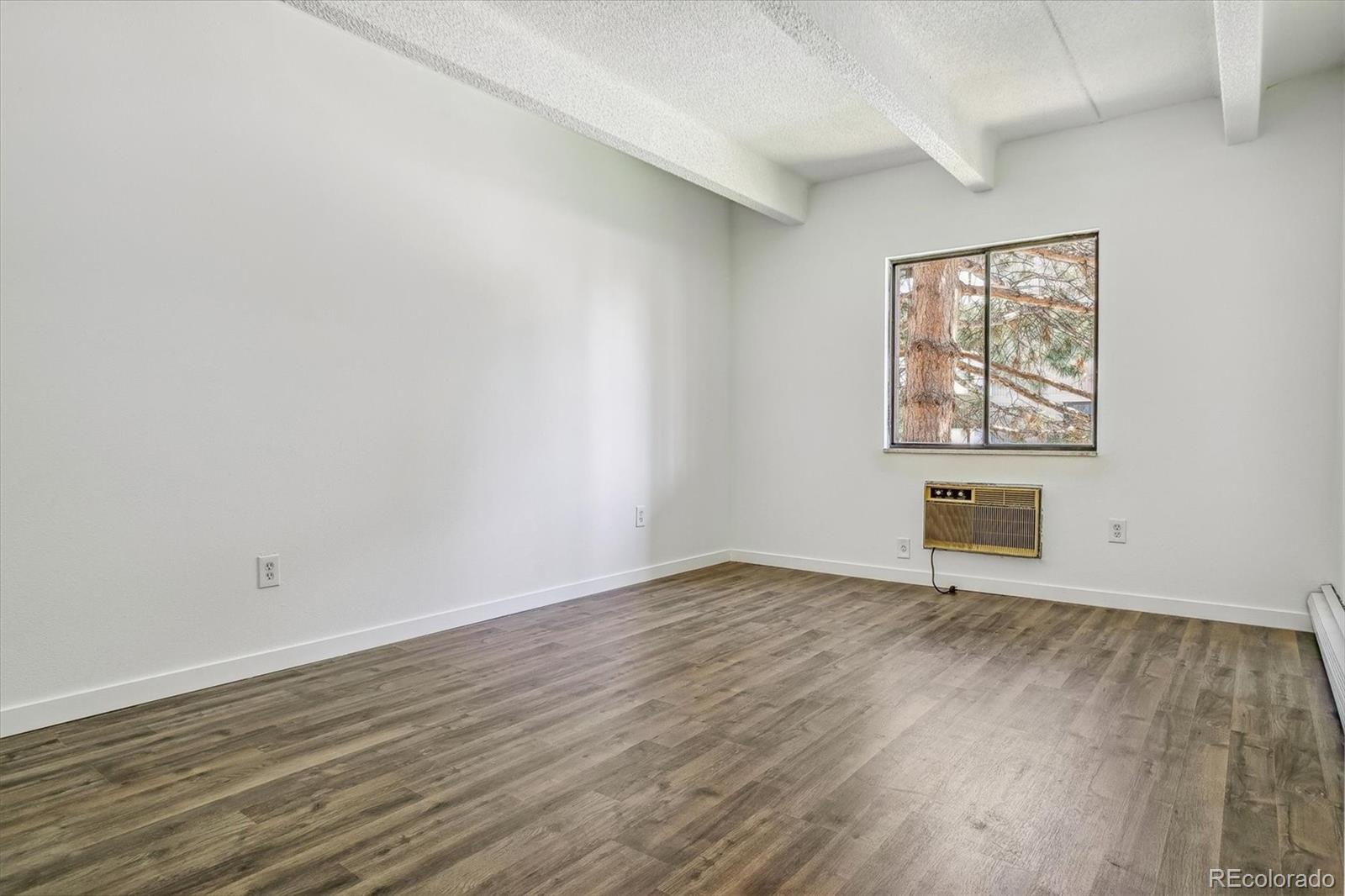 Empty room with plank flooring, exposed ceiling beams, and a window above a wall-mounted air conditioner.