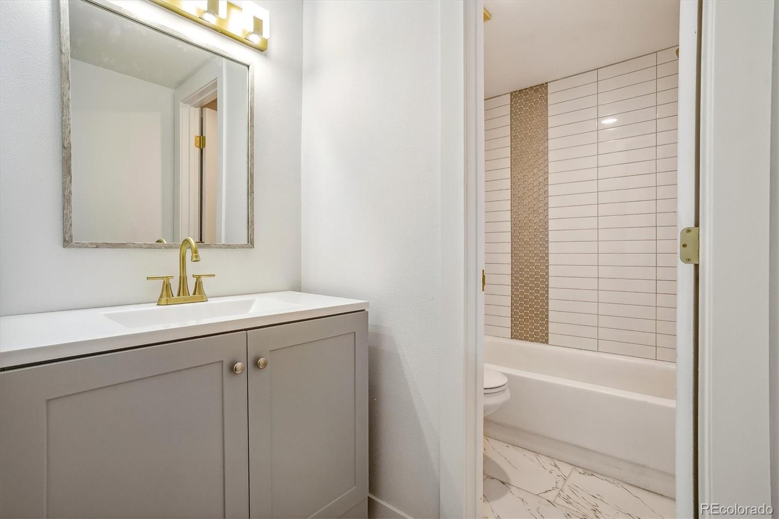 Bathroom with a gray vanity, brass faucet, and bathtub featuring a vertical mosaic tile accent.