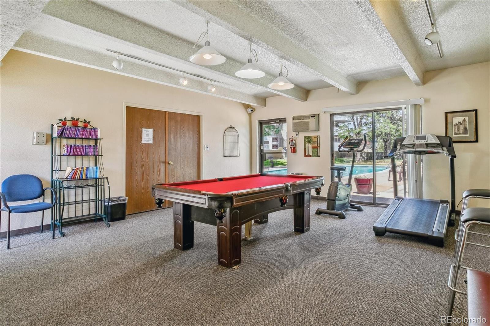 Community recreation room with a red-felt pool table, treadmills, and sliding doors facing an outdoor pool.