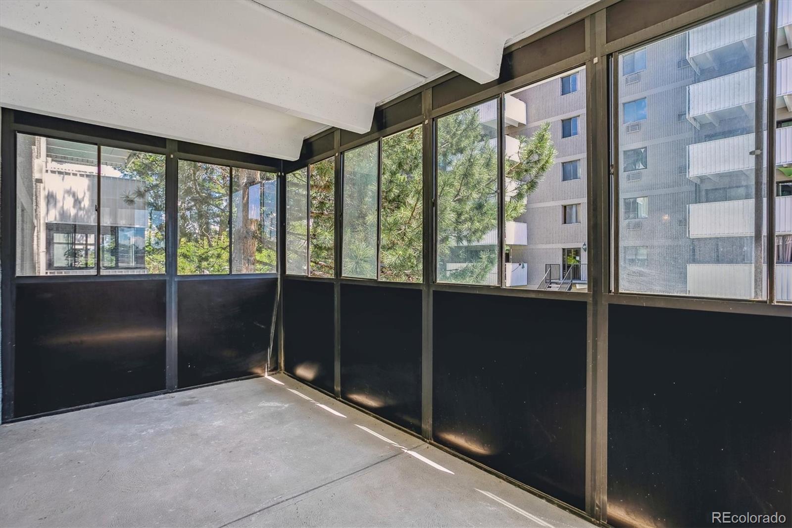 Enclosed balcony with floor-to-ceiling windows overlooking trees and a neighboring apartment building.