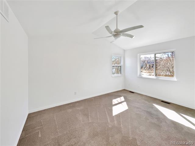 Empty bedroom with beige carpet, a ceiling fan, and two windows overlooking leafless trees.