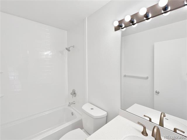 White bathroom with a tiled tub and shower, toilet, and vanity with mirror and lights.