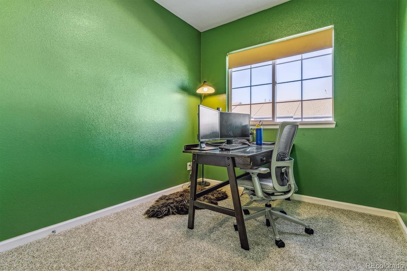 Home office with green walls, a desk with two monitors, an ergonomic chair, and a large window.