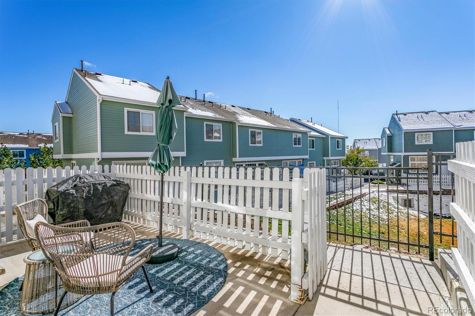 Fenced patio with woven chairs and a covered grill, facing teal townhomes with snow-covered roofs.