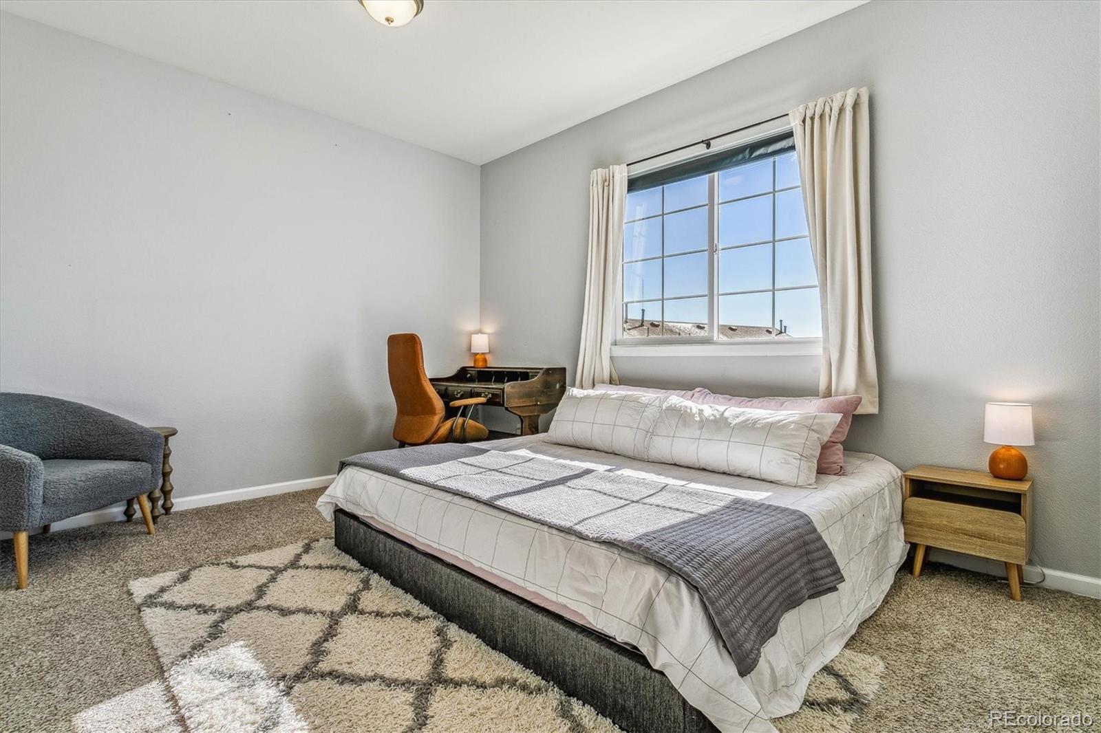 Staged bedroom with a large bed, desk by a bright window, and patterned area rug.