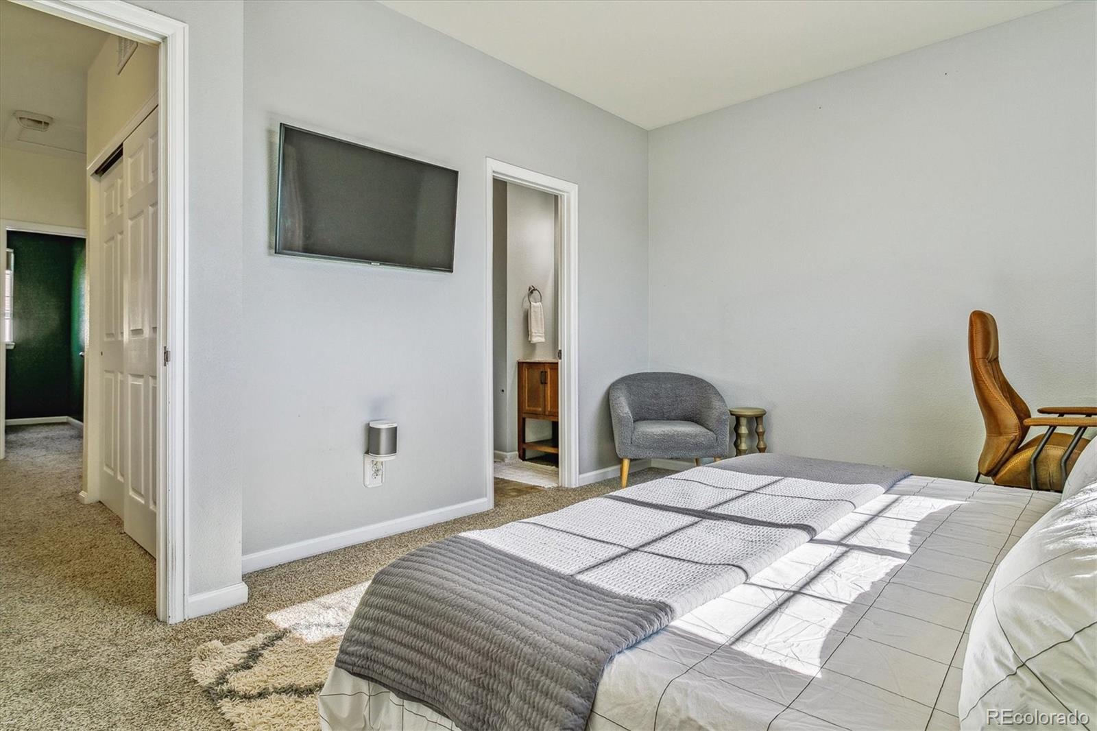 Bedroom with white bedding and a gray quilt, wall-mounted television, carpet, and gray accent chair.