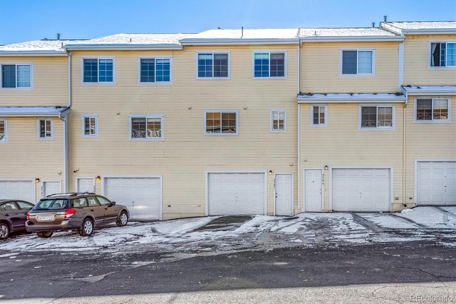 Beige attached townhome with garage doors and a snow-covered parking area.