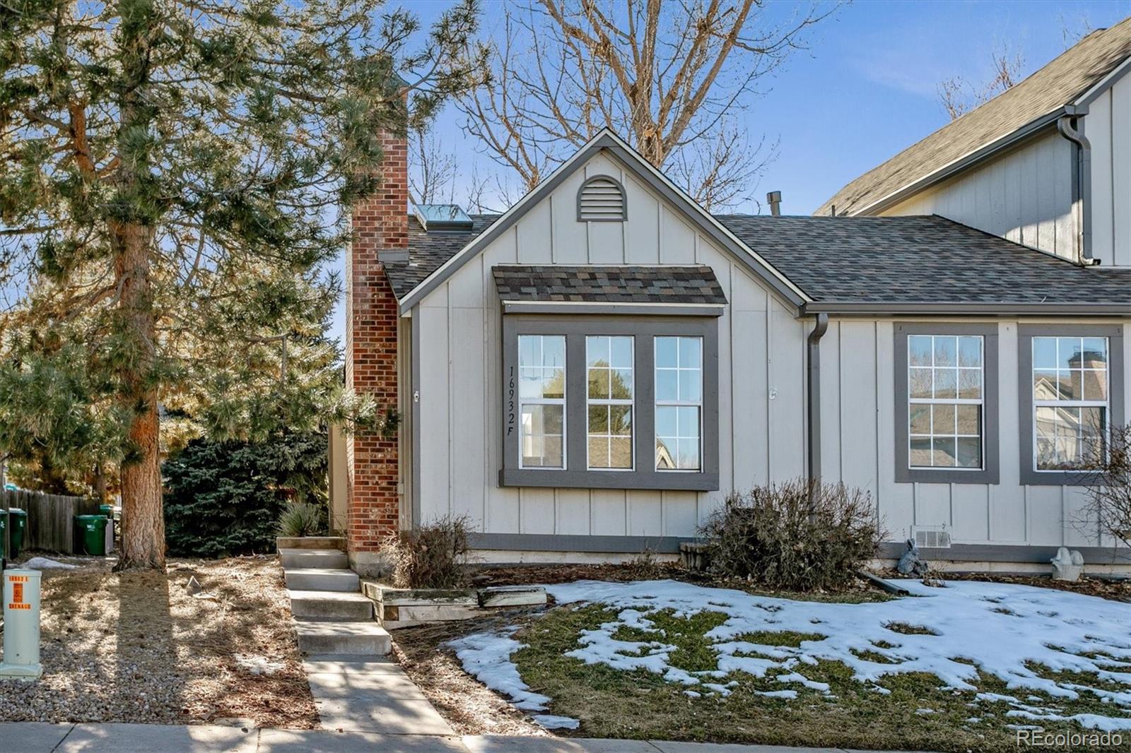 Light-gray townhouse exterior with a peaked entry gable, brick chimney, and snow patches in the yard.