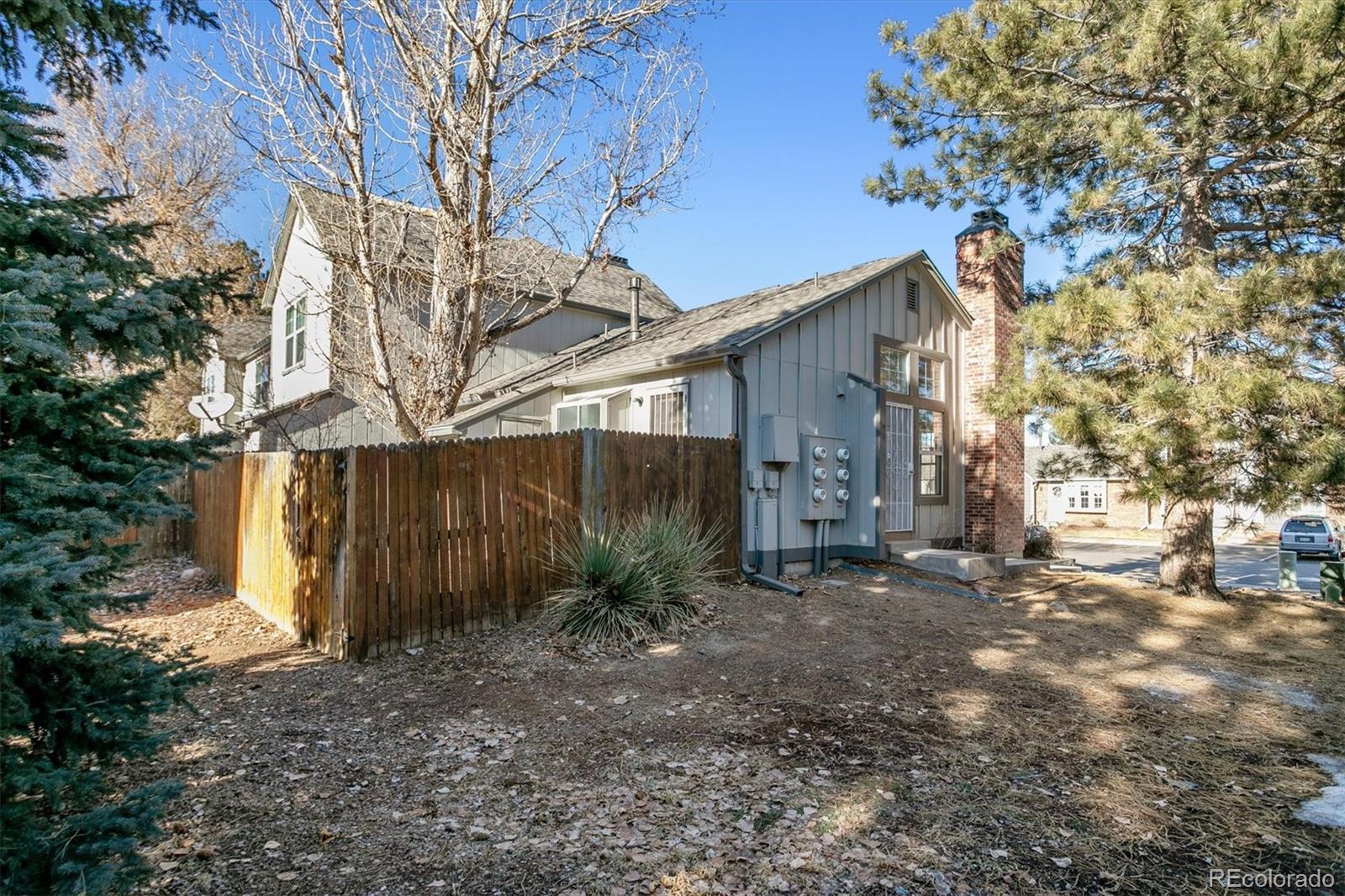 Gray townhouse side with brick chimney, wooden fence, and mature trees.