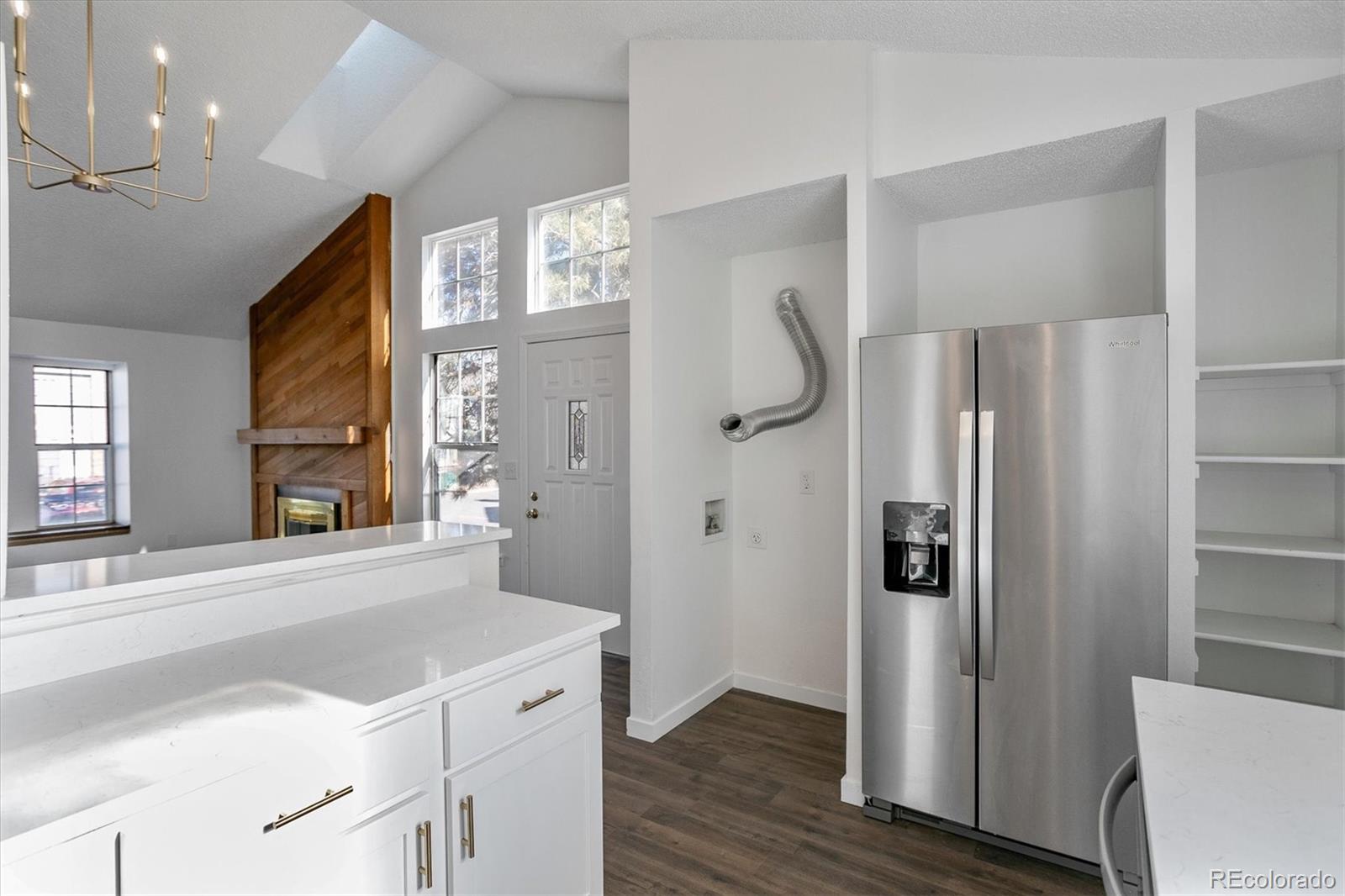 Bright kitchen with white counters and stainless steel refrigerator, opening to a vaulted living area with a wood fireplace.