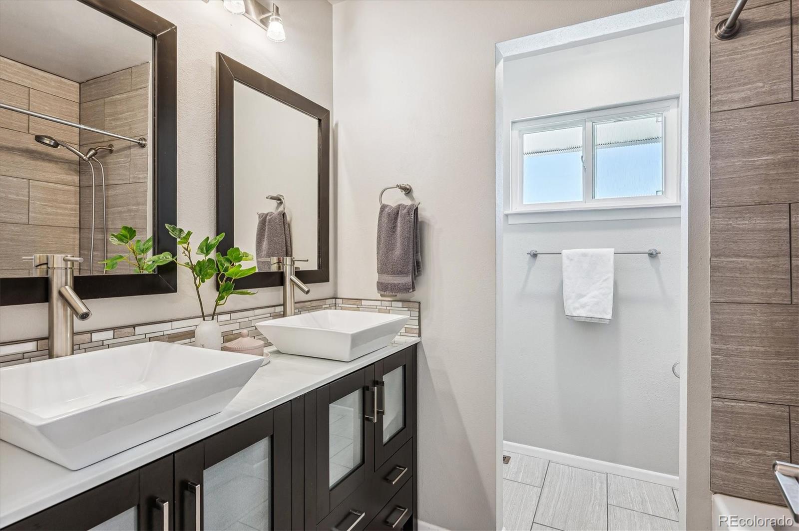 Bathroom with a dark double vanity, two vessel sinks, and a window above the shower area.