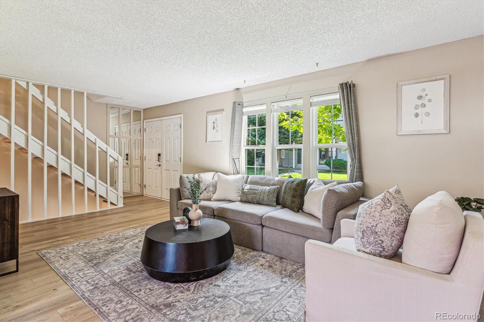 Staged living room with a gray sectional, round black coffee table, and staircase beside the entry.