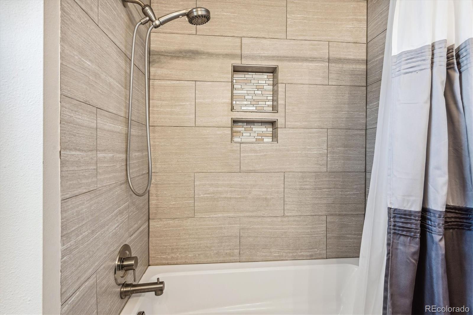 Bathtub and shower with beige tile, two recessed niches, handheld showerhead, and striped curtain.