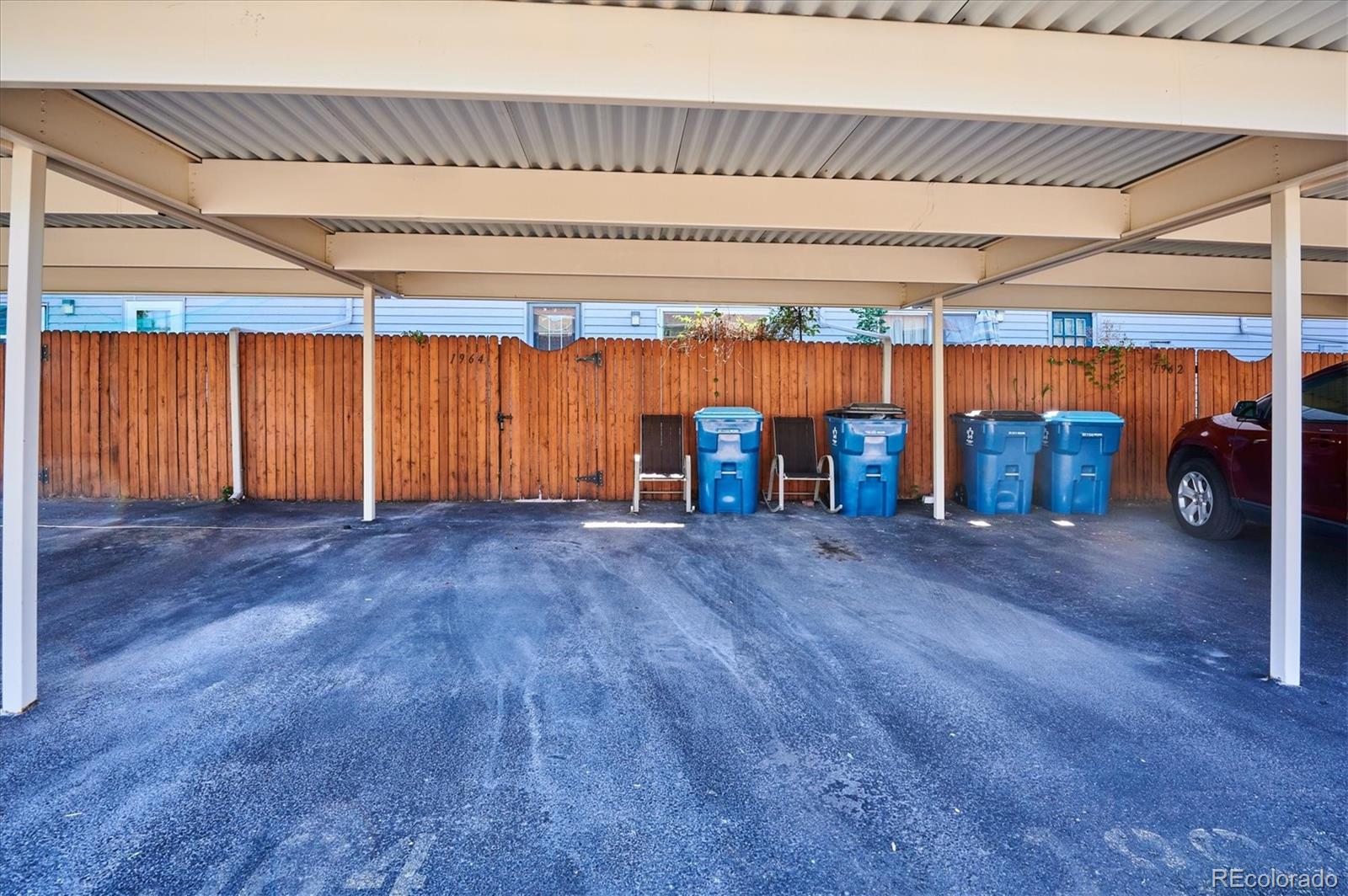 Covered carport with an asphalt parking area, wooden fence, and blue bins.