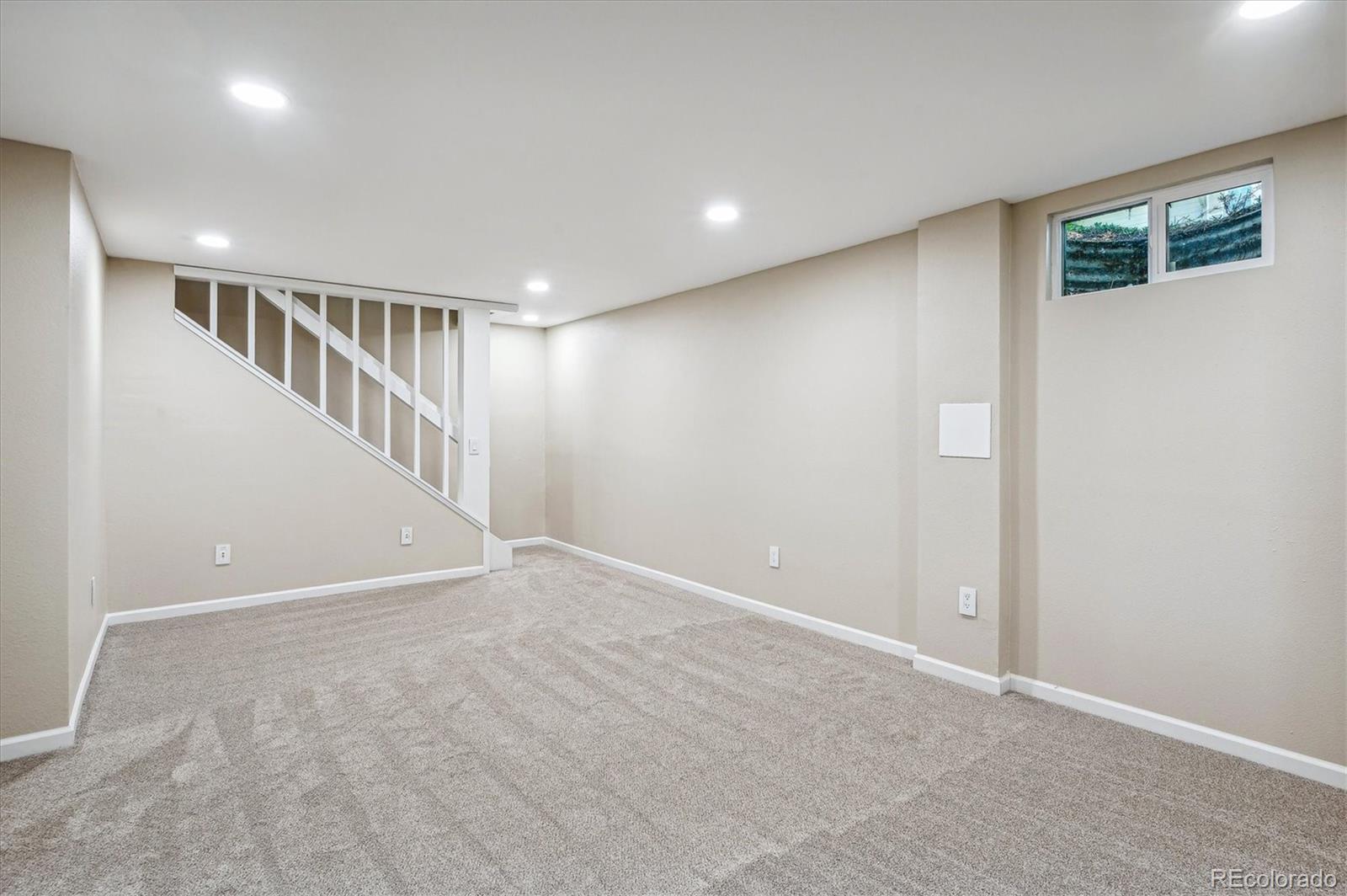 Vacant carpeted basement room with recessed lights, a small high window, and stairs with white railing.