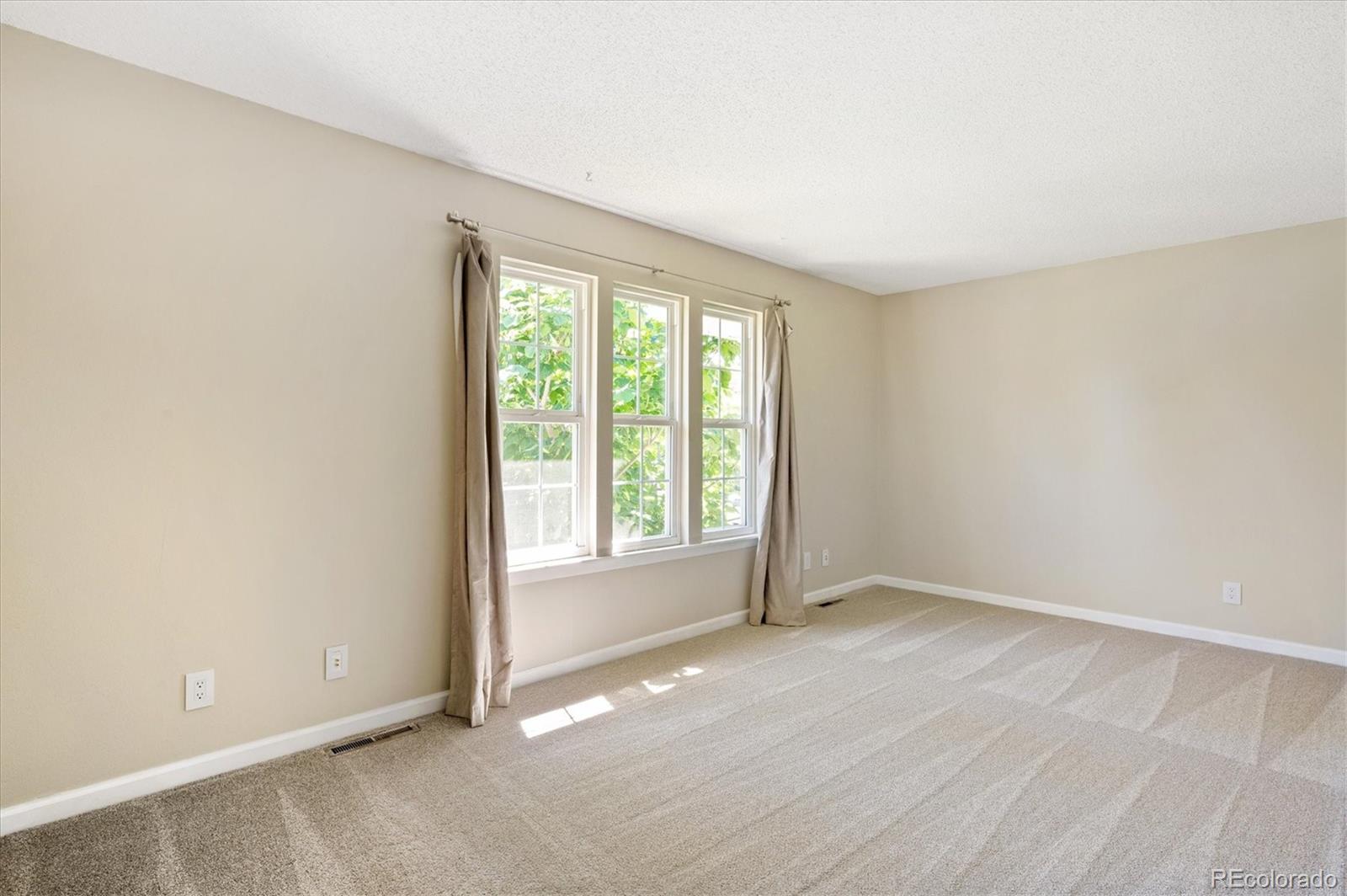 Empty carpeted room with three curtained windows and sunlight across the floor.