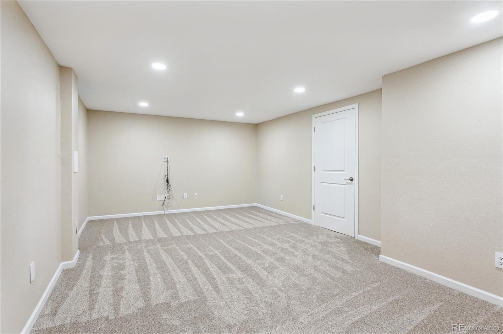 Empty finished room with gray patterned carpet, recessed lights, and a white paneled door.