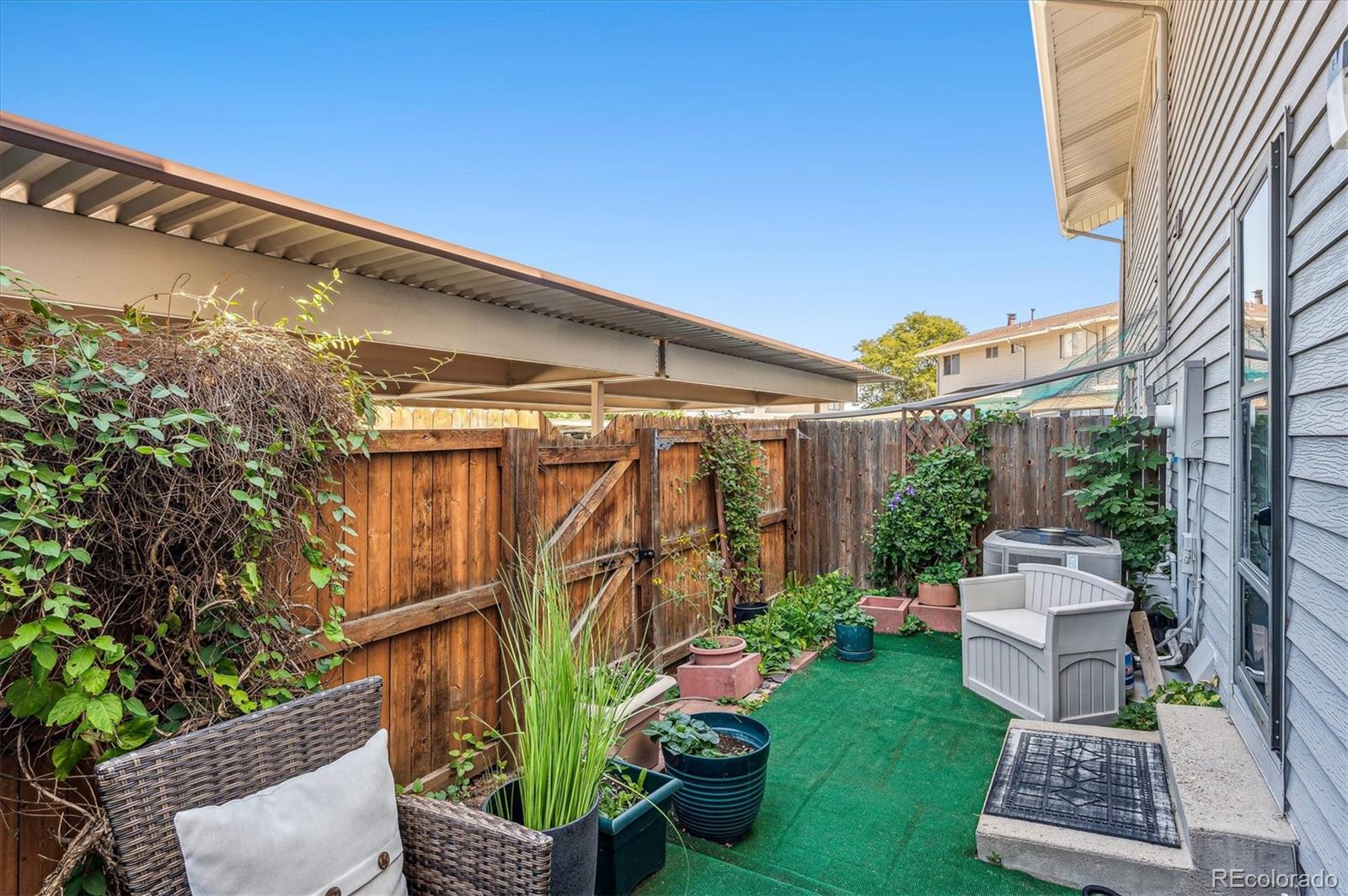 Fenced patio with artificial turf, garden planters, outdoor seating, and a covered carport.