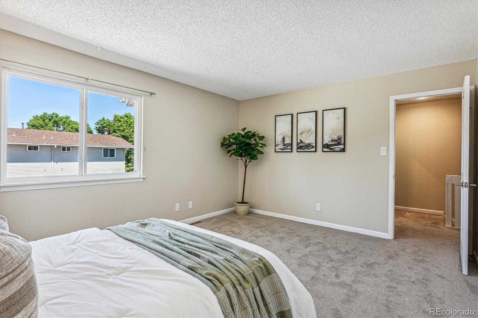 Staged bedroom with a large window, beige carpet, and a bed dressed in white and sage green.