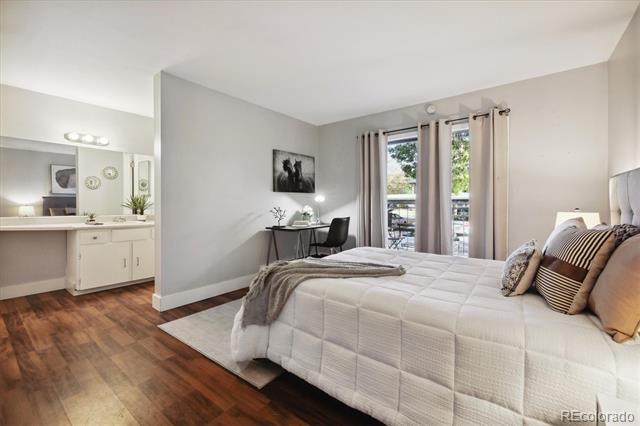 Bedroom with a white quilted bed, wood floors, vanity, and desk beside balcony doors.