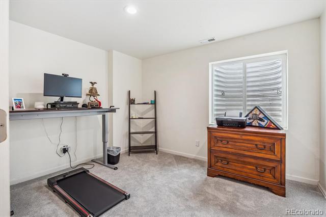 Carpeted room with a treadmill, desk and monitor, and wooden dresser beneath a shuttered window.