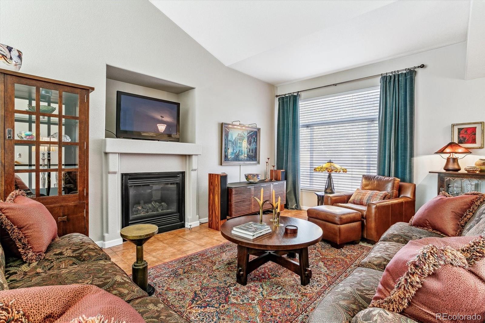 Living room with a gas fireplace, vaulted ceiling, and teal curtains framing a large window.