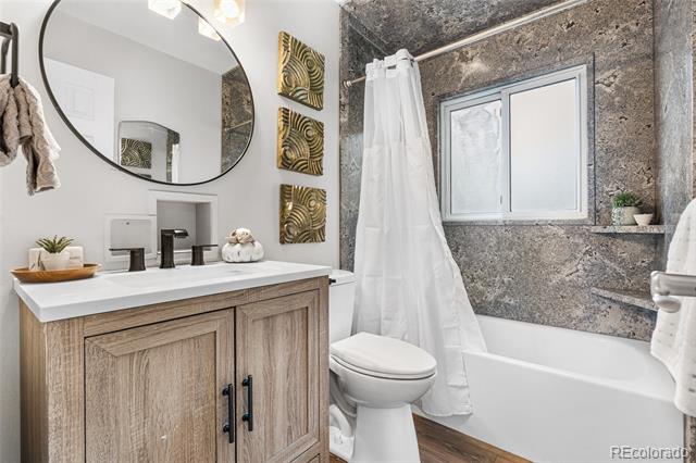 Bathroom with a wood vanity, round mirror, white tub and gray stone-look tile surround.