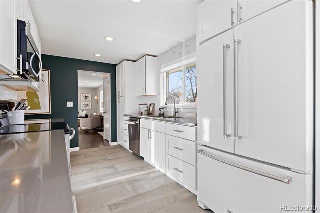 Bright kitchen with white cabinetry, a large refrigerator, gray counters and a window overlooking trees.